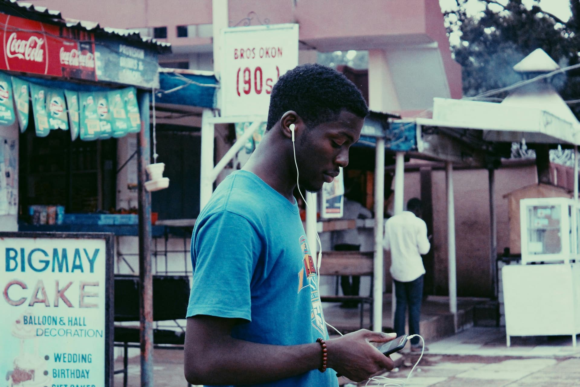 A young man in an African city street listens to his phone with earbuds.